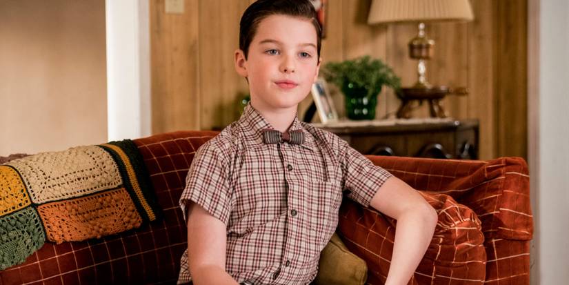Young Sheldon smiles as he sits and leans against the side of the couch with one arm
