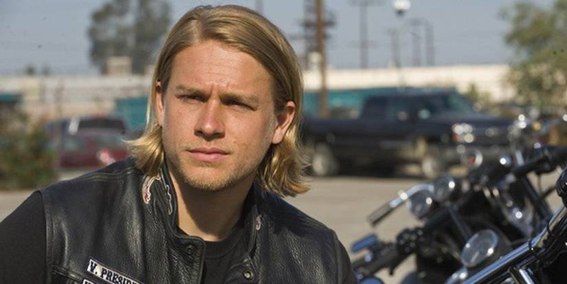 A solemn Jax Teller with a leather jacket stands by a motorbike in a car park