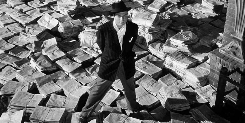 Charles Foster Kane stands with his hands behind his back surrounded by newspapers.