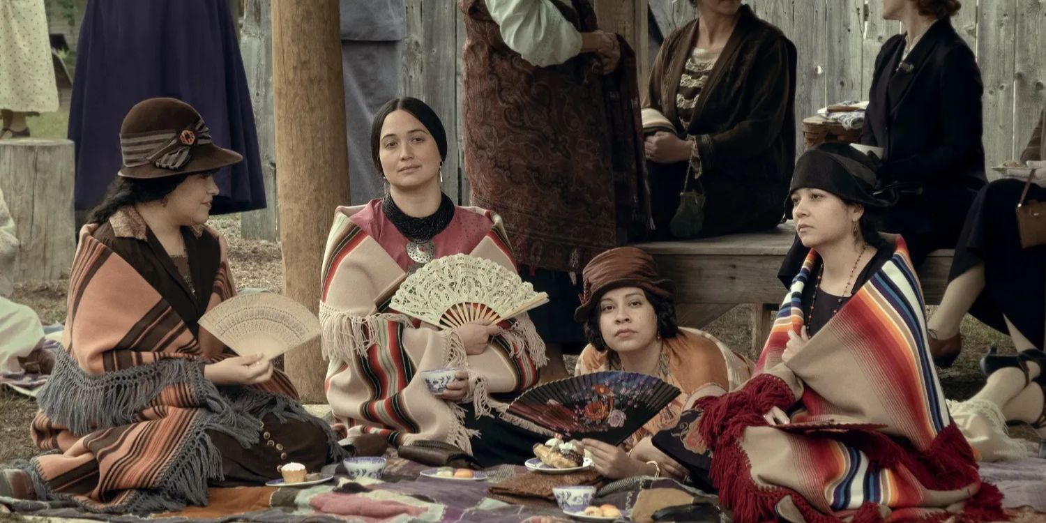 Mollie de Lily Gladstone segura um leque e está sentada com outras mulheres em Assassinos da Lua das Flores