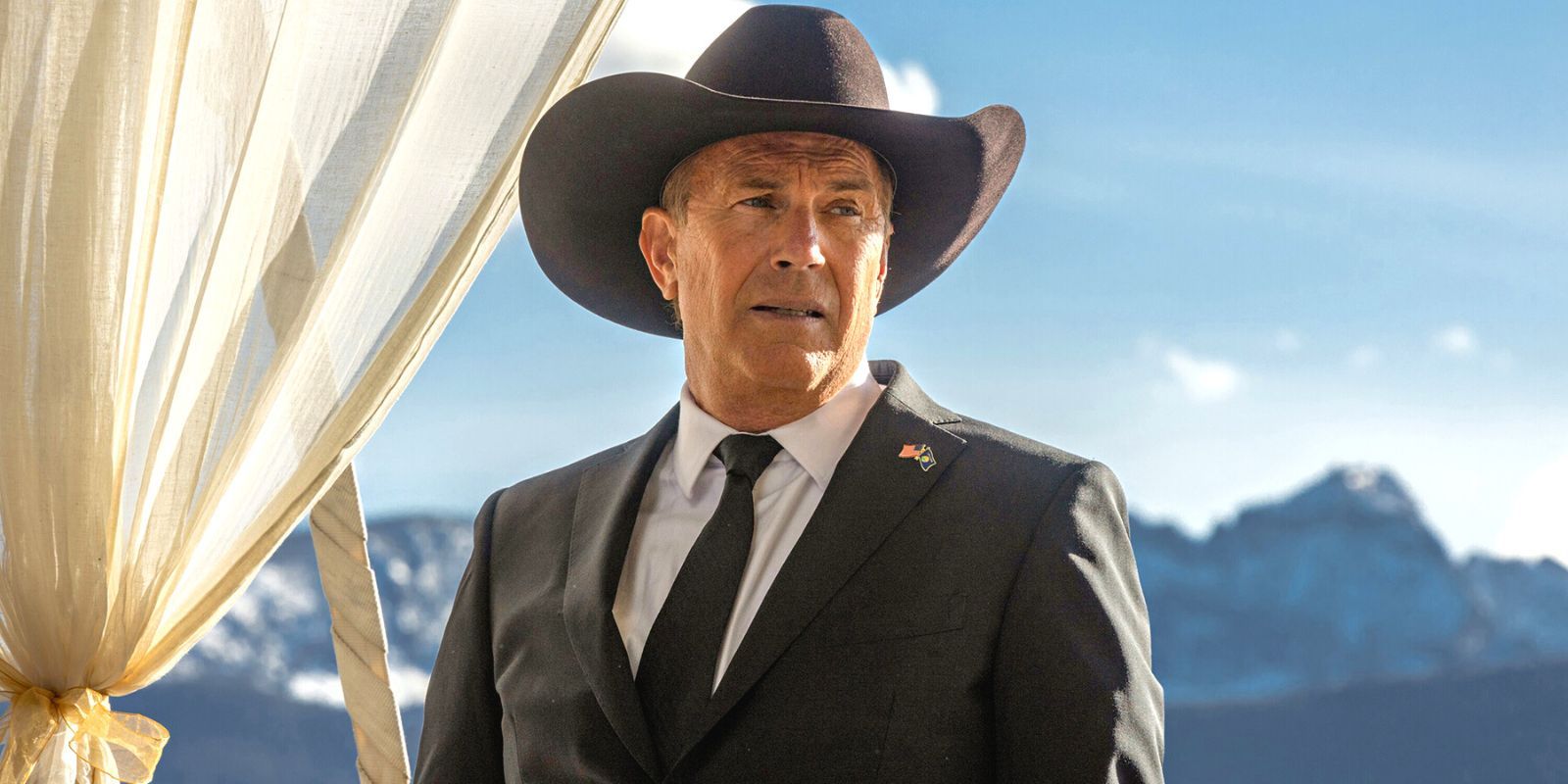 Kevin Costner in Yellowstone in a black suit and hat stands in front of a mountain range