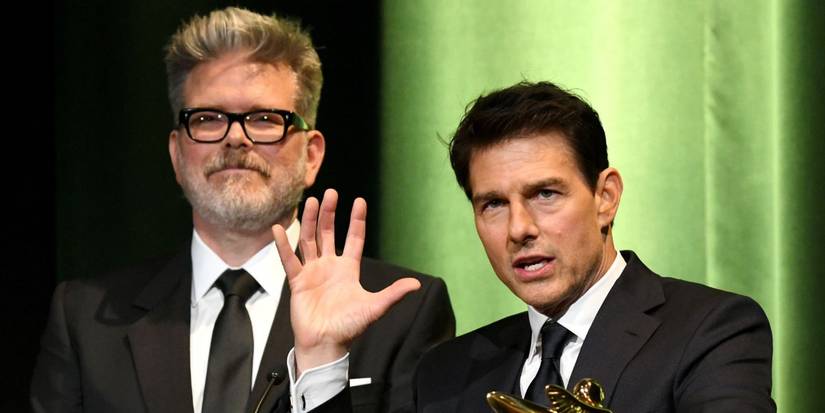 Director Christopher McQuarrie and actor Tom Cruise, both wearing black suits, stand at a podium