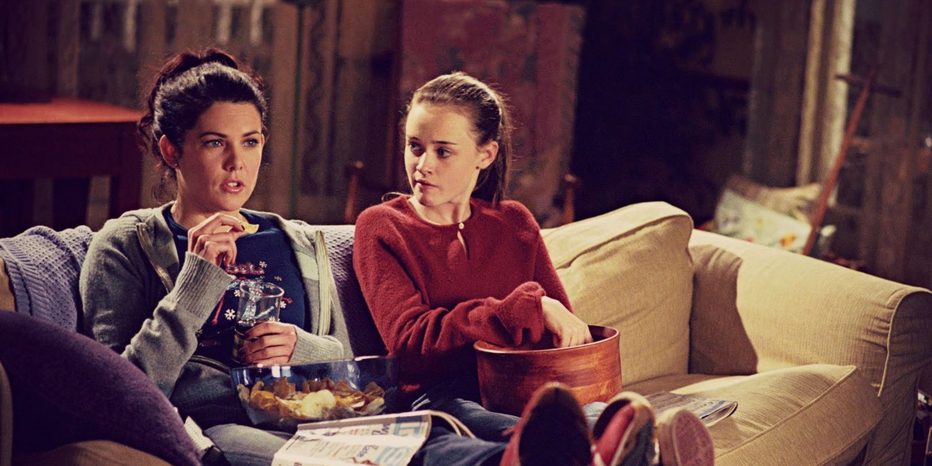Rory and Lorelai Gilmore sit on the couch and eat popcorn in Gilmore Girls.