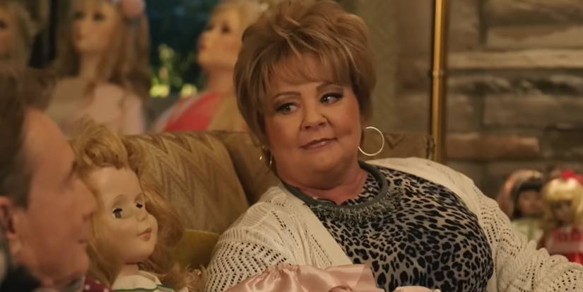 Charles' sister (Melissa McCarthy) talks to Oliver while sitting on a couch with dolls in Only Murders in the Building.
