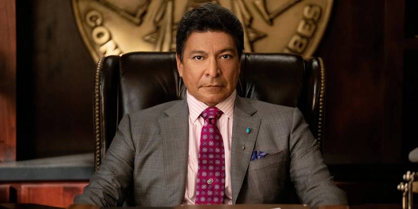 Thomas Rainwater, played by Gil Birmingham, is wearing a grey suit and is seated at his desk in Yellowstone.