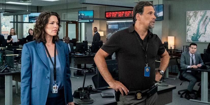 Isobel, in blue suit, stands next to Jubal in a brown shirt with hands on hips in the office in FBI