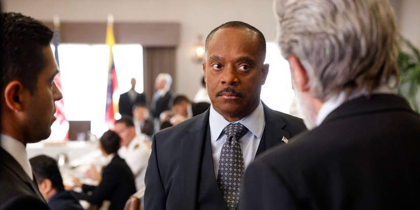 Leon Vance wears a suit and looks at Agents Parker and Torres, who are back to camera, in NCIS