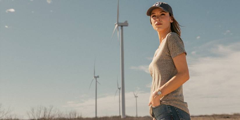 Rebecca, played by Kayla Wallace, wearing a T-shirt and ballcap next to wind turbines in Landman