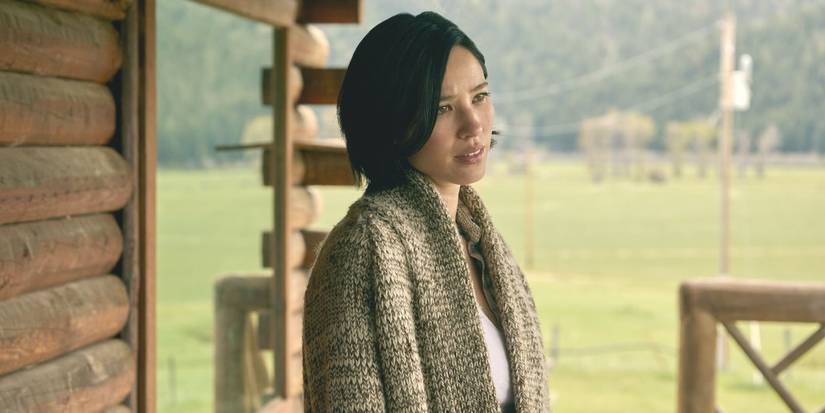 Monica Dutton, played by actor Kelsey Asbille, in a grey sweater on a cabin porch in Yellowstone