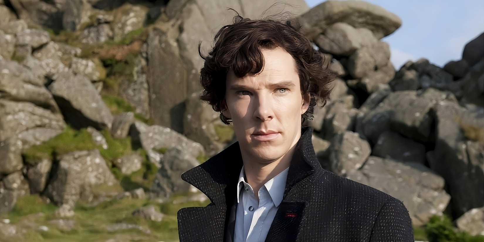 Benedict Cumberbatch wearing a black coat and standing in front of a cliff as Sherlock Holmes