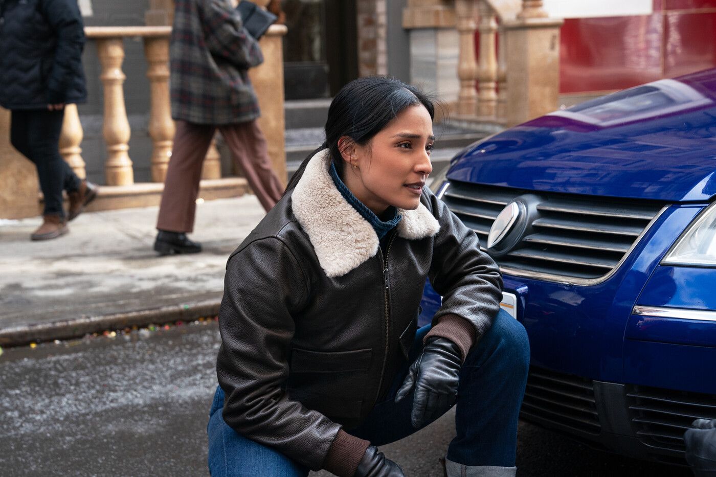 Dani Rhodes, played by Emily Alabi, wearing a bomber jacket and kneeling next to a blue car in FBI