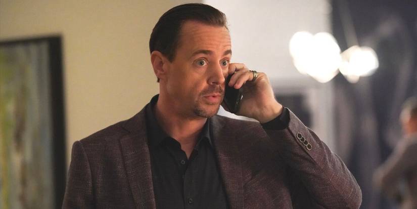 Timothy McGee wearing a brown jacket and black shirt, looking surprised as he holds a phone in NCIS