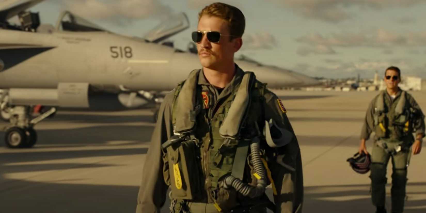 Rooster, wearing full flight suit, walks past fighter jets in front of Maverick in Top Gun Maverick