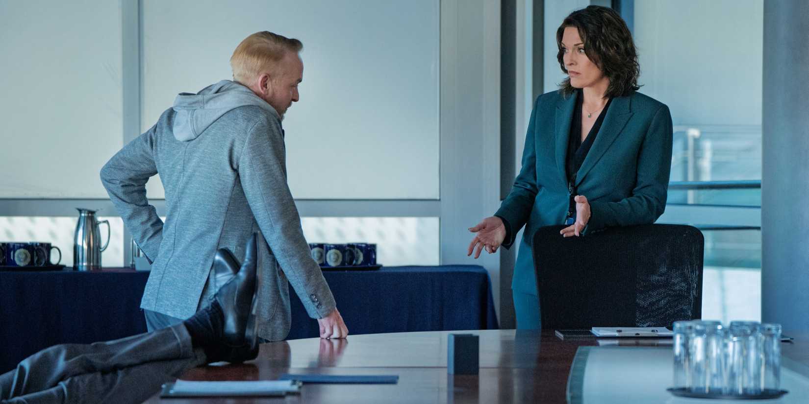 Isobel, in a blue suit, gestures to Scott, back to camera in a grey hoodie, next to a table on FBI