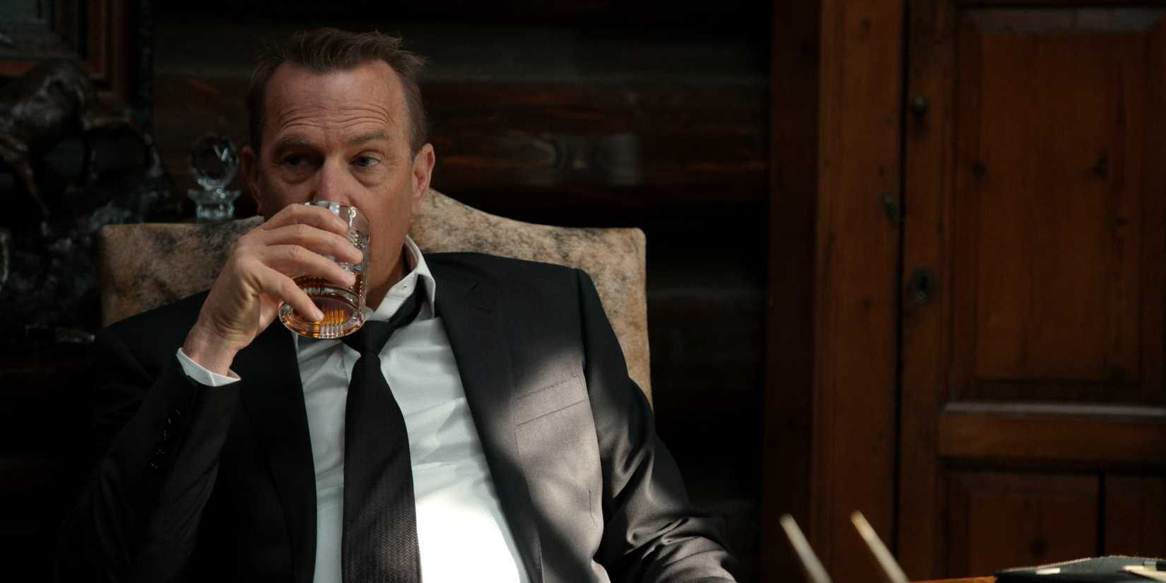 John Dutton, wearing a suit, drinks whiskey in Yellowstone.