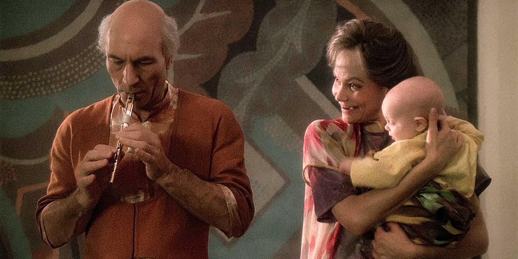 Jean Luc PIcard as Elderly Kamin playing his flute in an orange outfit as Eline stands next to him holding a baby from Star Trek The Next Generation
