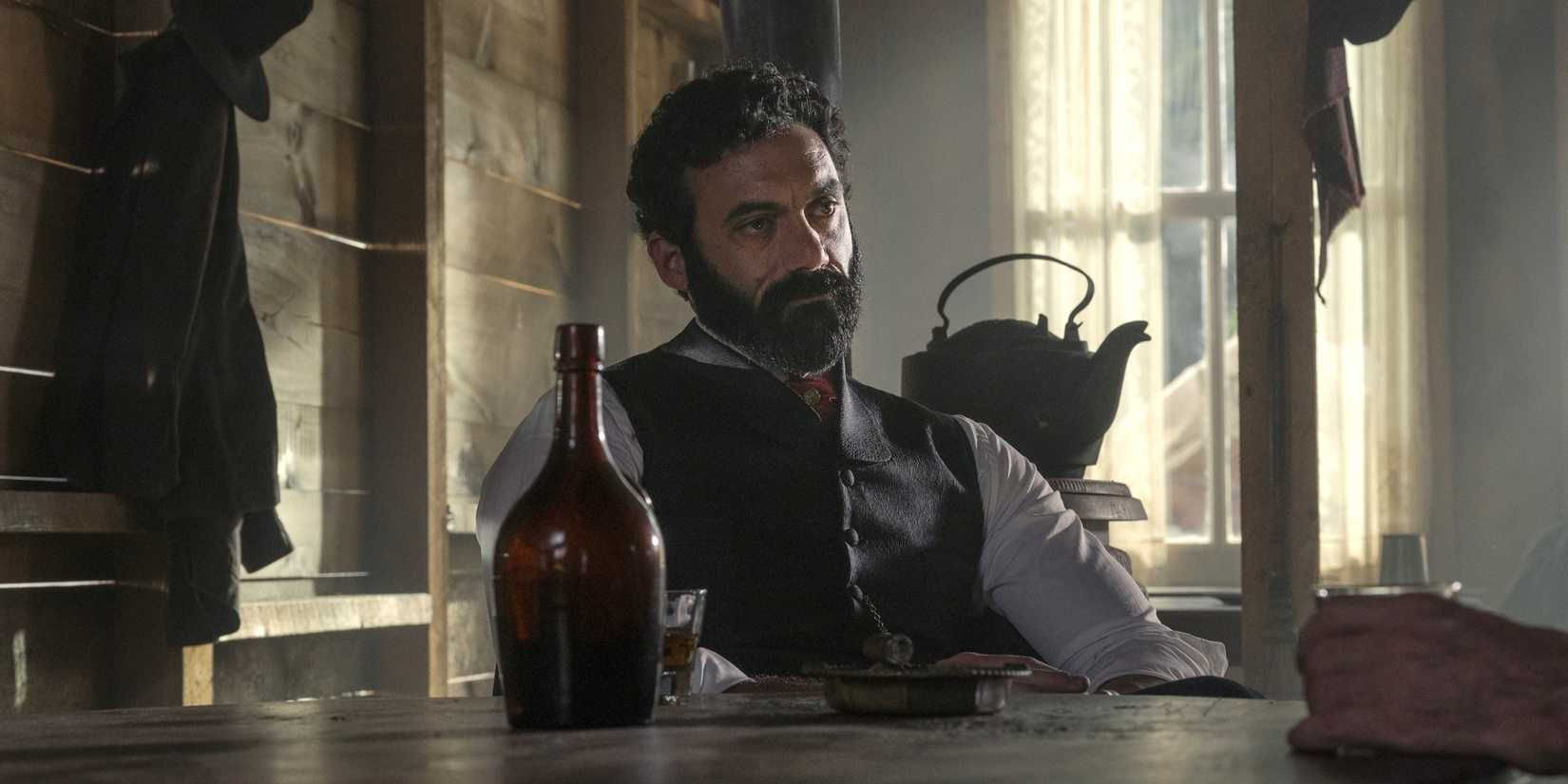 George Russell (Morgan Spector) sitting in at a table with a bottle in a saloon in Morenci in The Gilded Age Season 3.