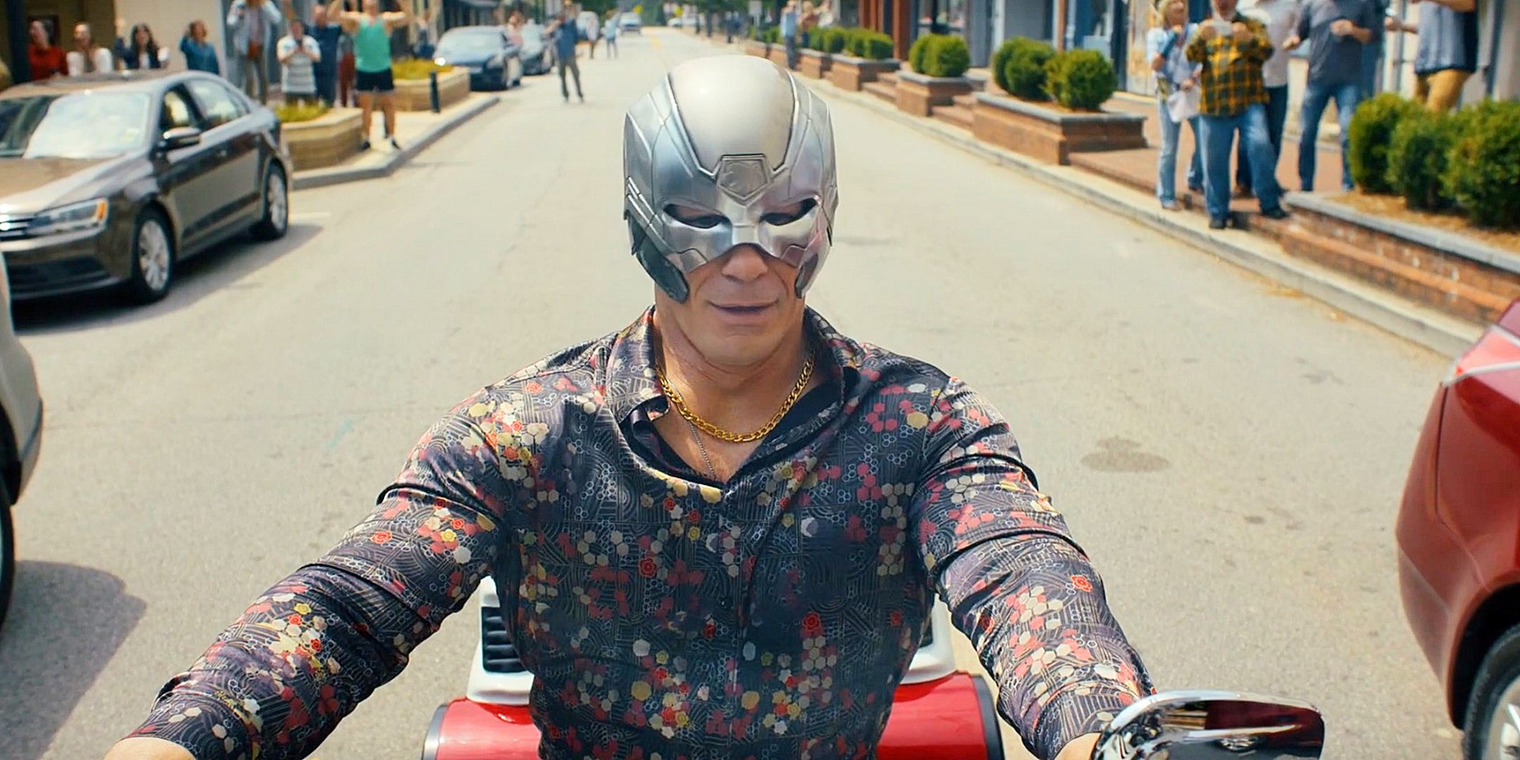 Peacemaker (John Cena) on his motorcycle with townspeople behind him from Peacemaker 2