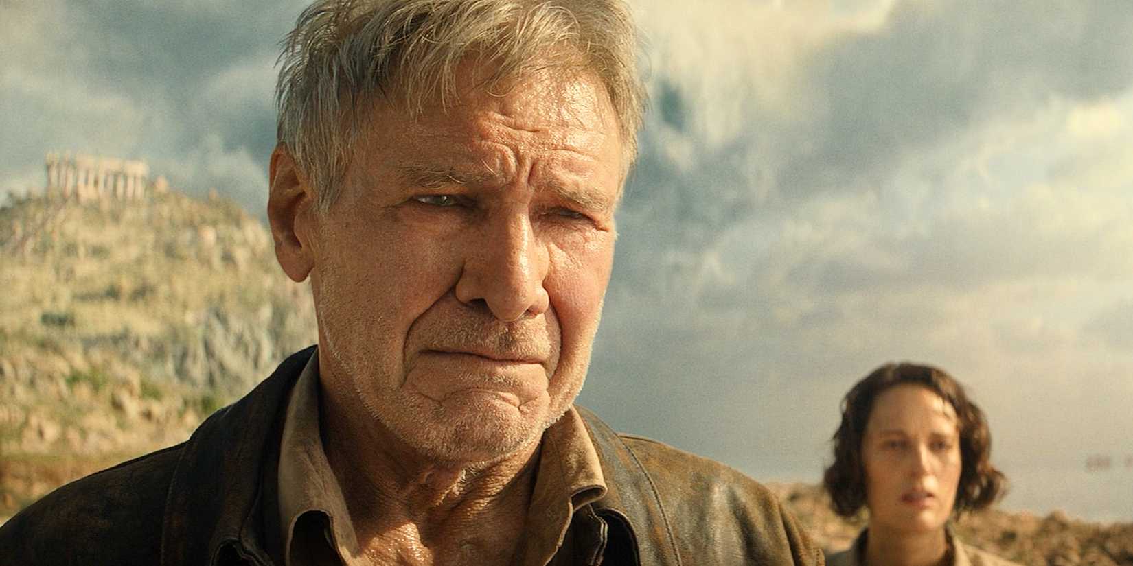 Close up of a sad Indiana Jones with ancient greek ruins and a blue, cloudy sky behind him from the Dial of Destiny
