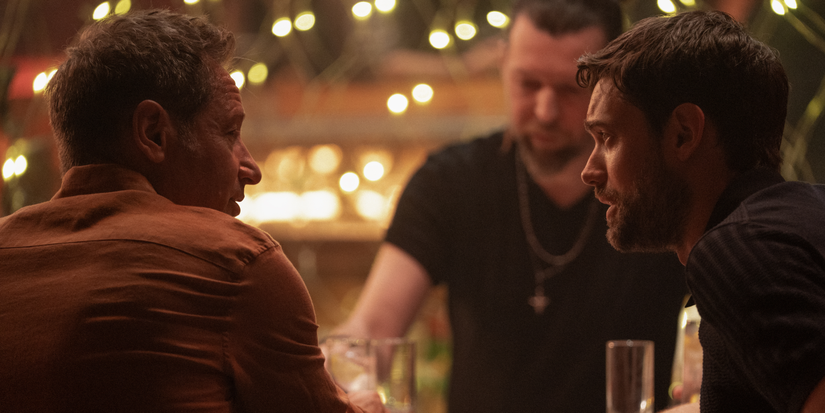 Adam and Jamie sitting at a club bar talking in Malice on Prime Video