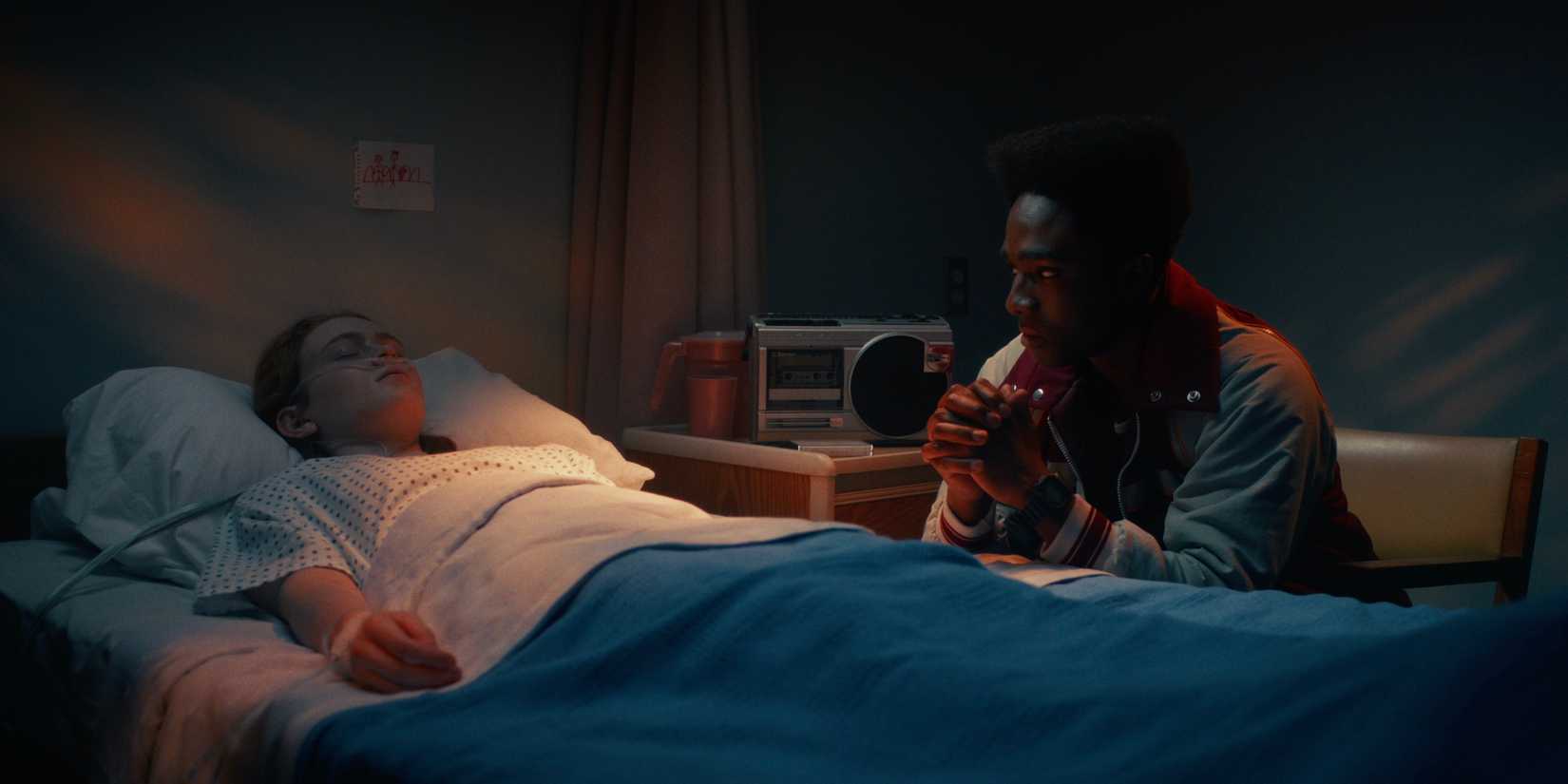 Lucas (Caleb McLaughlin) sitting next to Max (Sadie Sink), who's lying in a hospital bed, on Stranger Things