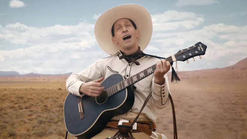 Tim Blake Nelson singing and playing guitar as a cowboy in the desert