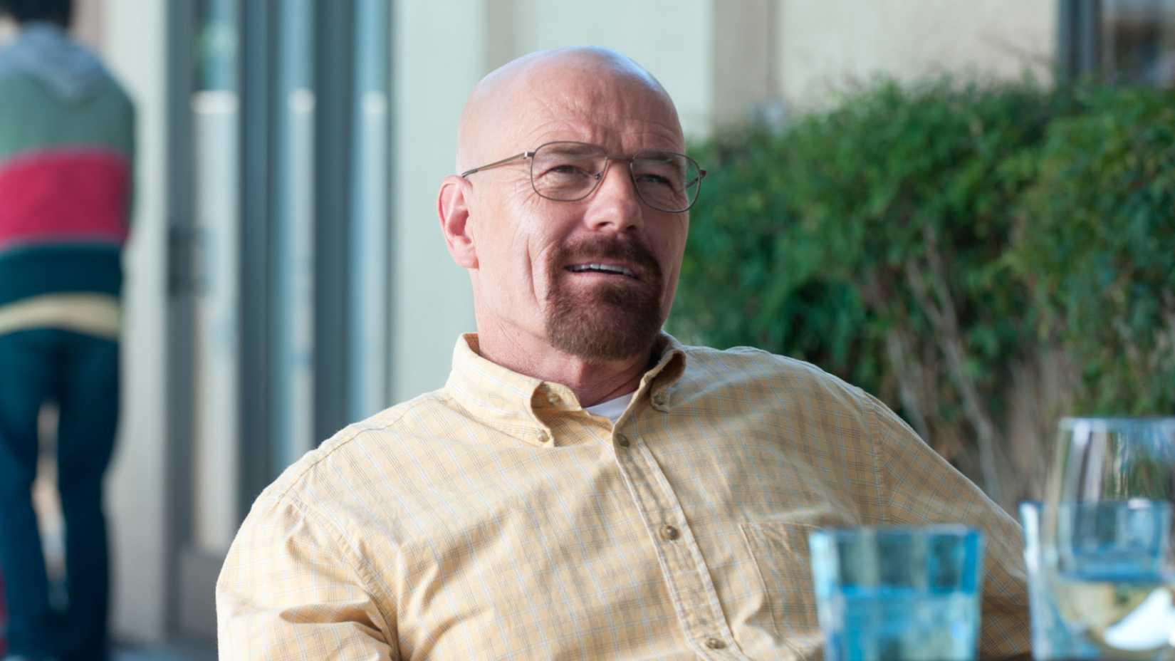 Walter White sits at an outdoor dining table, smirking, in Breaking Bad