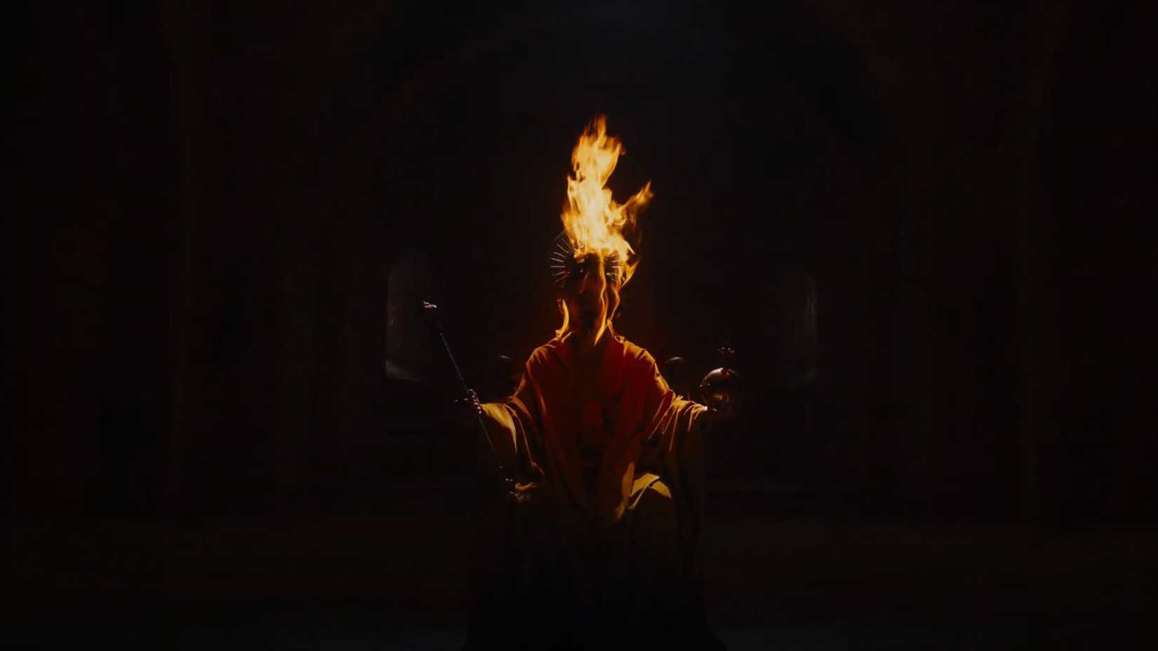 Dev Patel as Gawain sits on a throne in The Green Knight with fire coming from his head and crown. 