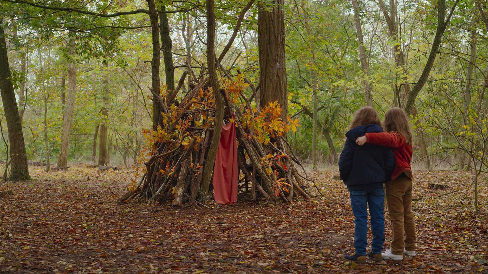 Two girls put their arms around each other as they look at their wood fort in a forest in Petite Maman.
