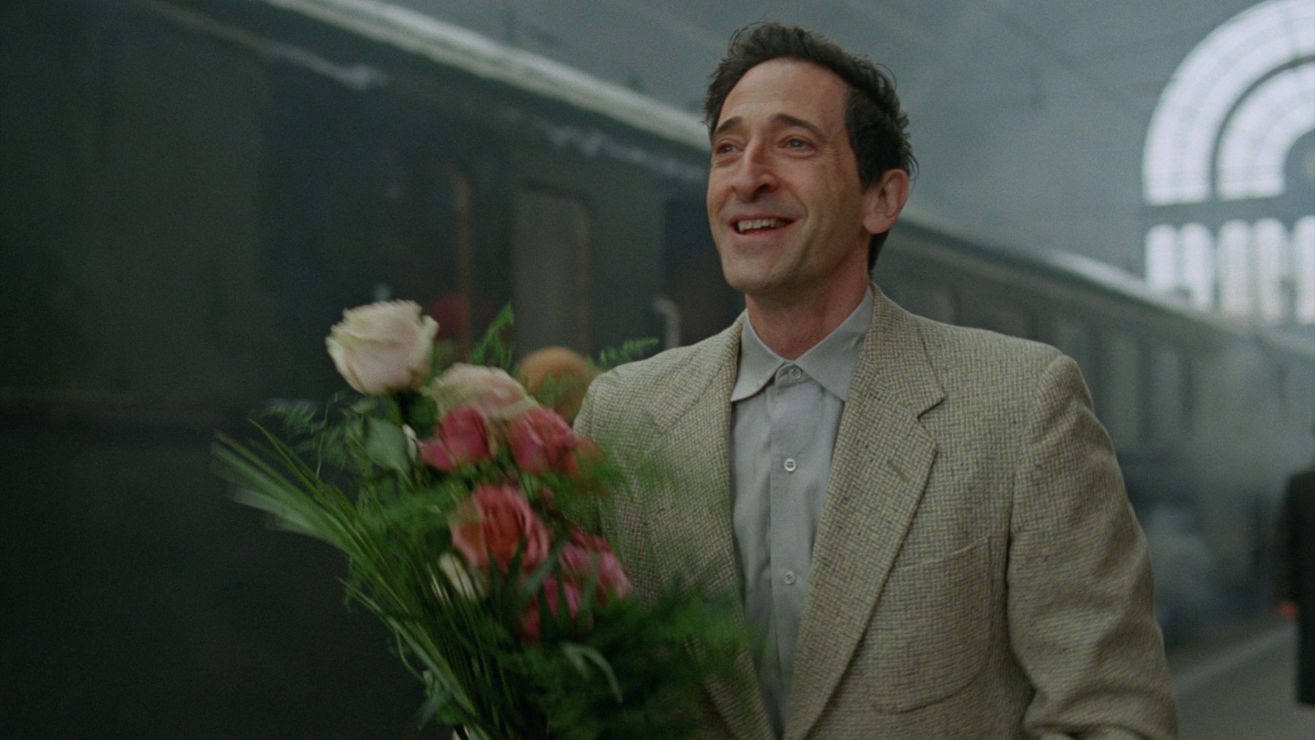 Adrien Brody holding a rose bouquet on a train station looking hopeful in The Brutalist